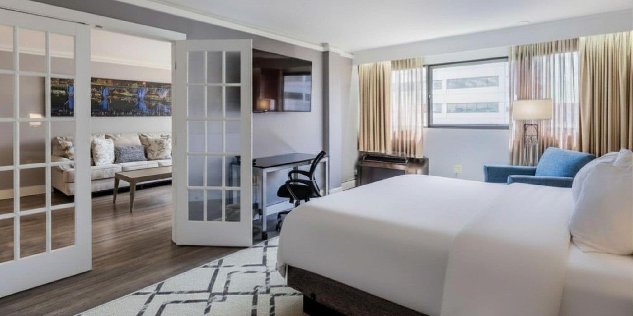A hotel room with a bed, a desk, a chair, a sofa, and a large window with curtains.