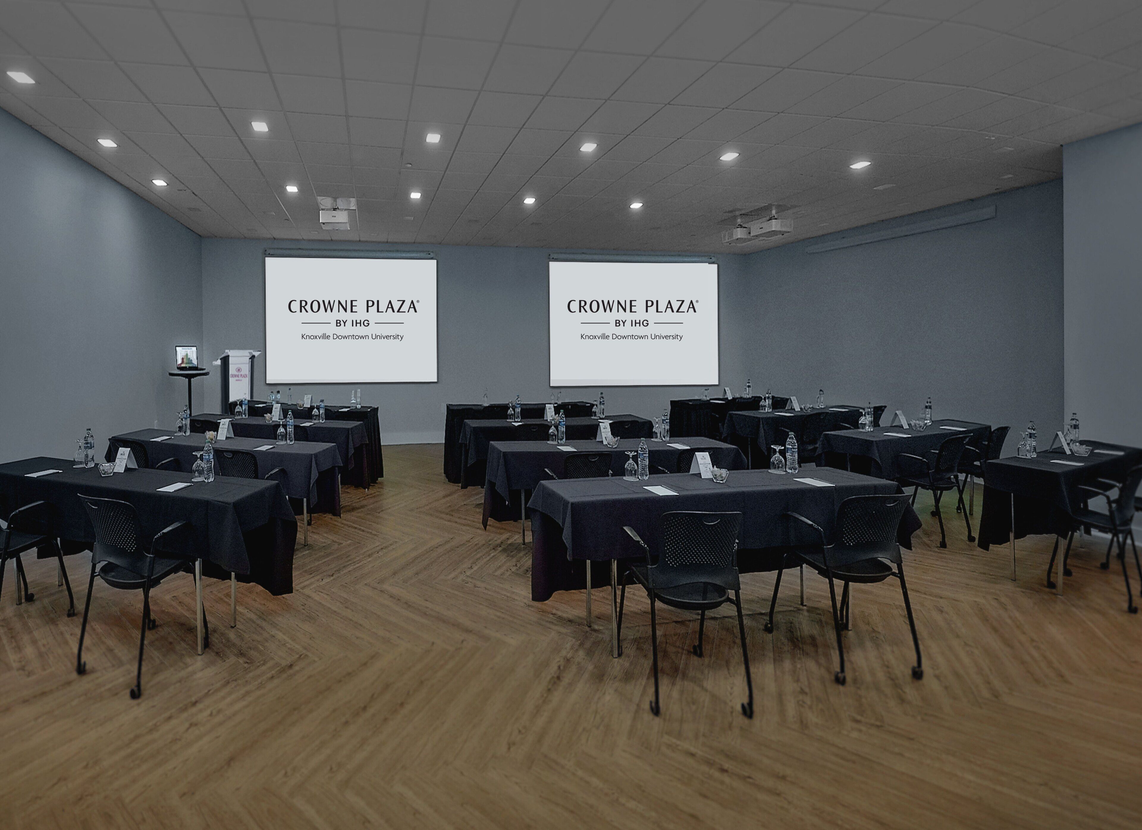A conference room with tables, chairs, and two screens displaying 'CROWNE PLAZA BY IHG' at Knoxville Downtown University.