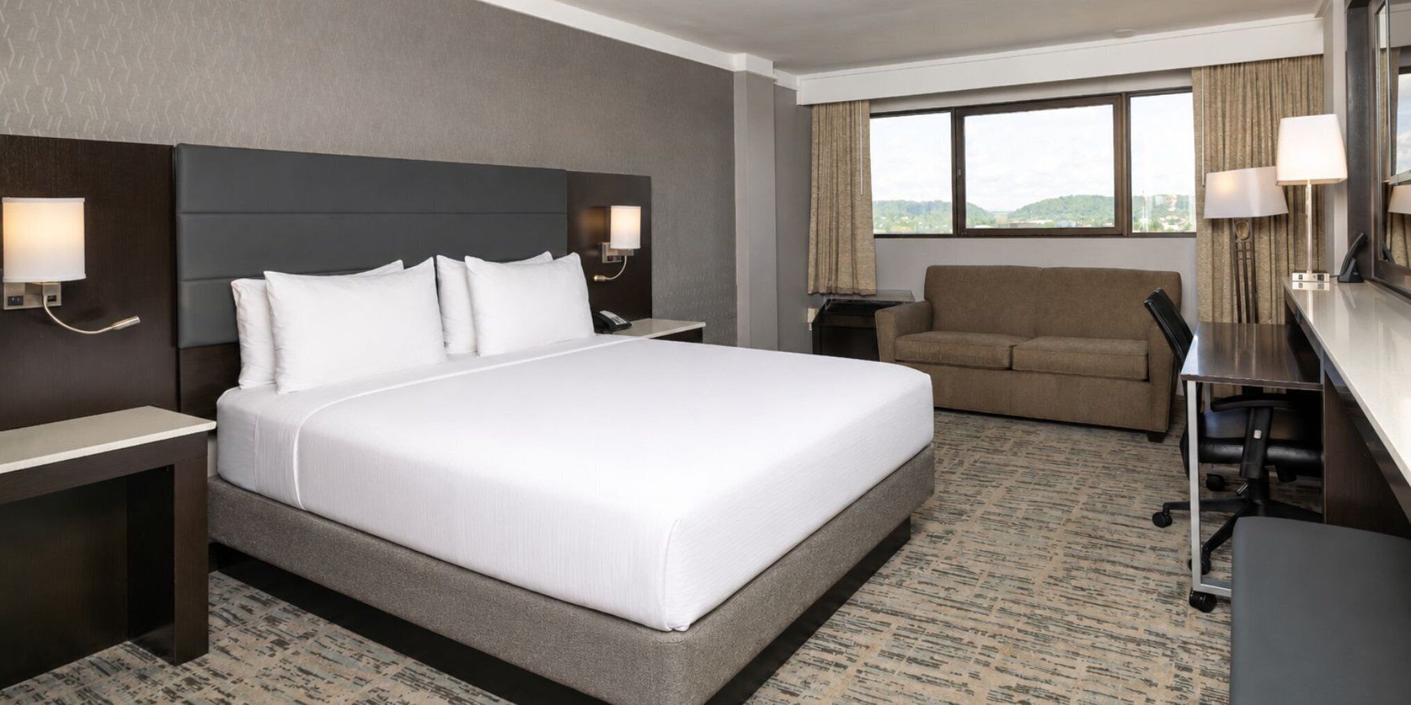 A hotel room with a large bed, a desk, a chair, a sofa, and a view of the outdoors.