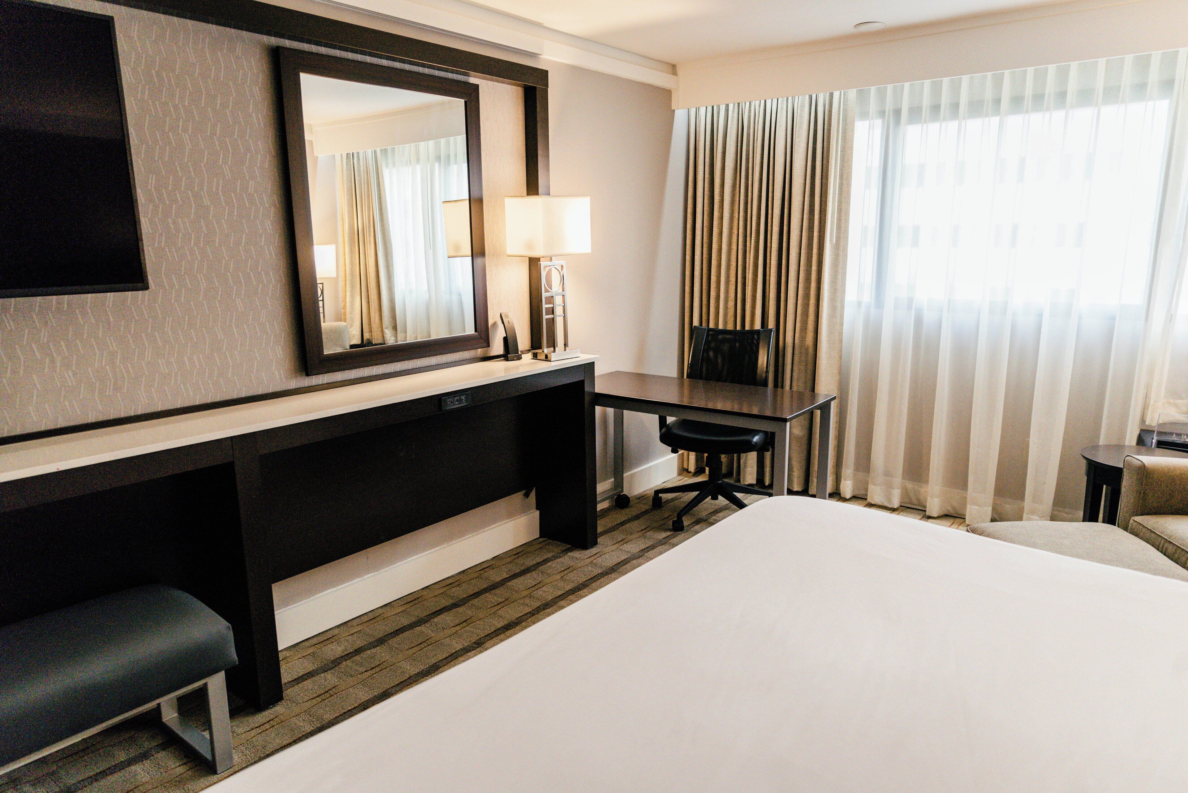 A hotel room with a large bed, a desk with a chair, a mirror, a lamp, and a view of the outside through sheer curtains.