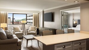 Moderni hotellihuone, jossa on sohvaryhmä, ruokapöytä, taulutelevisio ja näkymä kaupungin ylle.