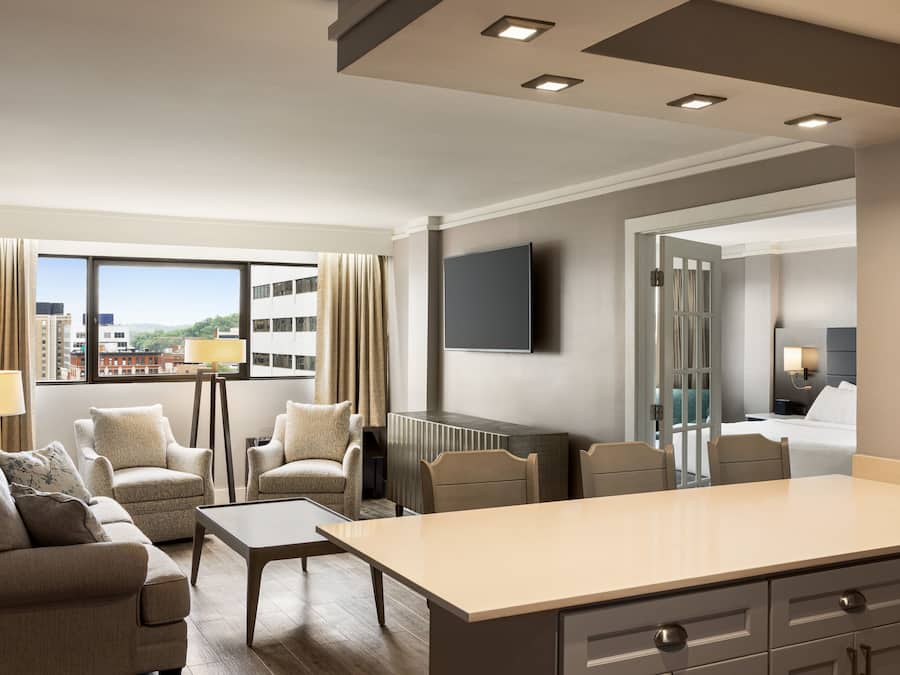 Ein modernes Hotelzimmer mit einer Sofagruppe, einem Esstisch, einem Flachbildfernseher und Blick auf die Stadtlandschaft.