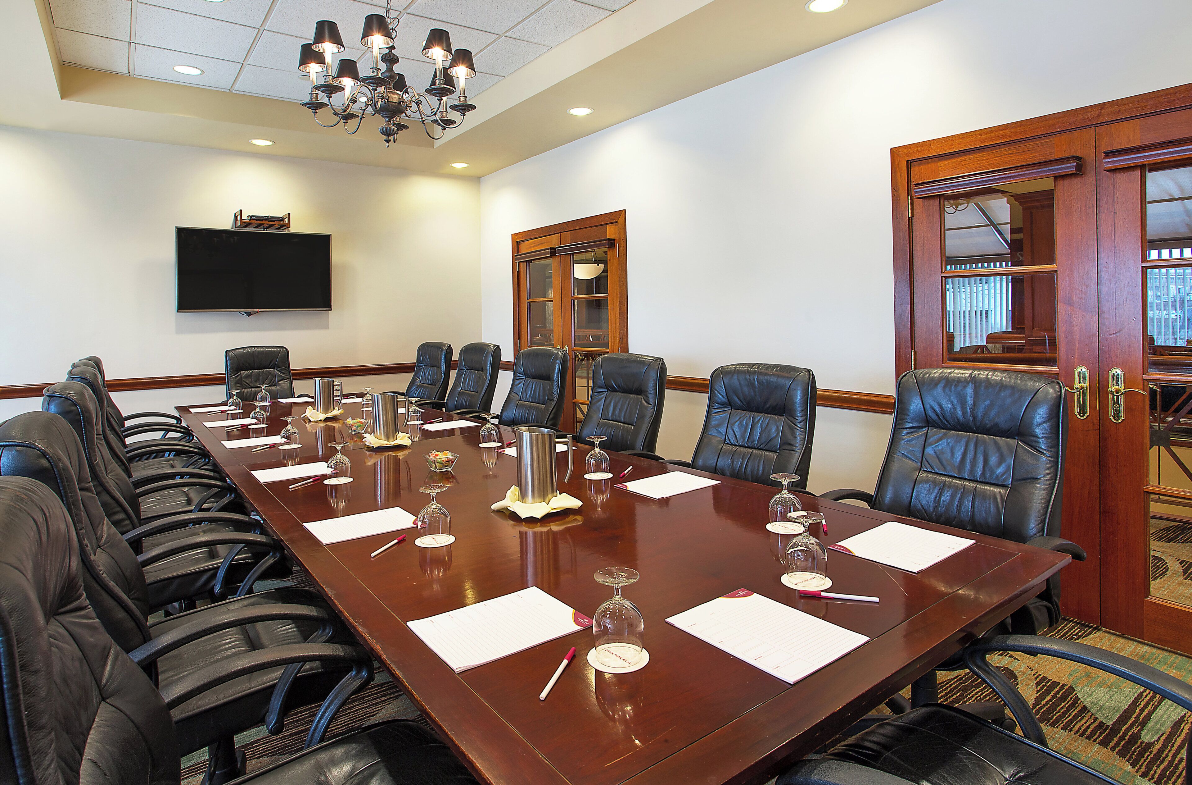 A conference room with a long table, multiple chairs, a flat-screen TV, and a chandelier.