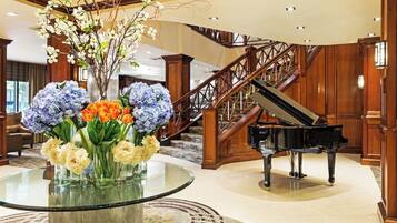 A grand lobby with a grand piano, a glass table with floral arrangements, and a staircase.