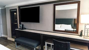 A hotel room with a flat-screen TV, a large mirror, a desk with a chair, a bench, and a small trash can.