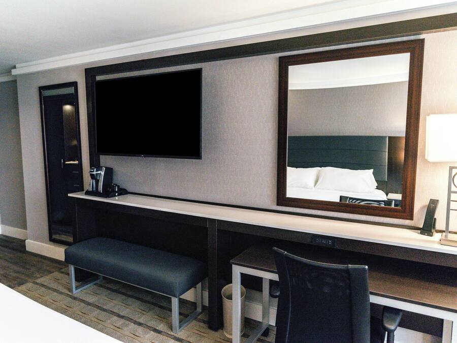 A hotel room with a flat-screen TV, a large mirror, a desk with a chair, a bench, and a small trash can.