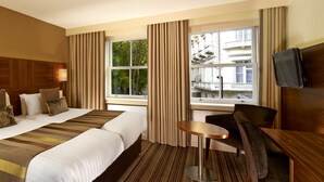A hotel room with two beds, a small round table, a chair, a TV, and a window with a view of buildings and trees. - Blakemore Hyde Park (London)