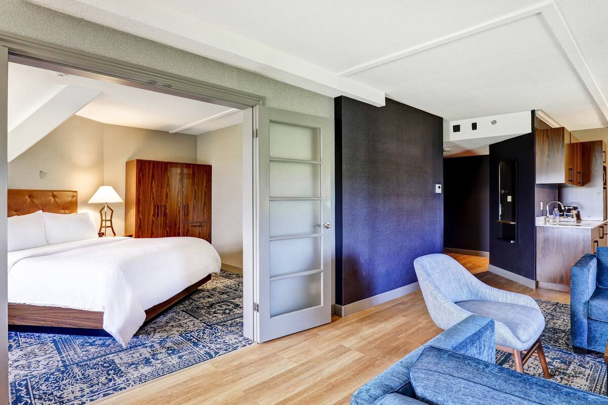 A modern hotel room with a large bed, a seating area with blue chairs, a wooden wardrobe, and a kitchen area visible through an open door.