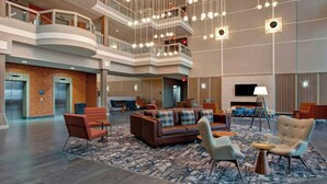 Un vestíbulo espacioso con un diseño moderno, que cuenta con una zona de estar central con sofás y sillones, una alfombra estampada y un techo alto con luces colgantes.