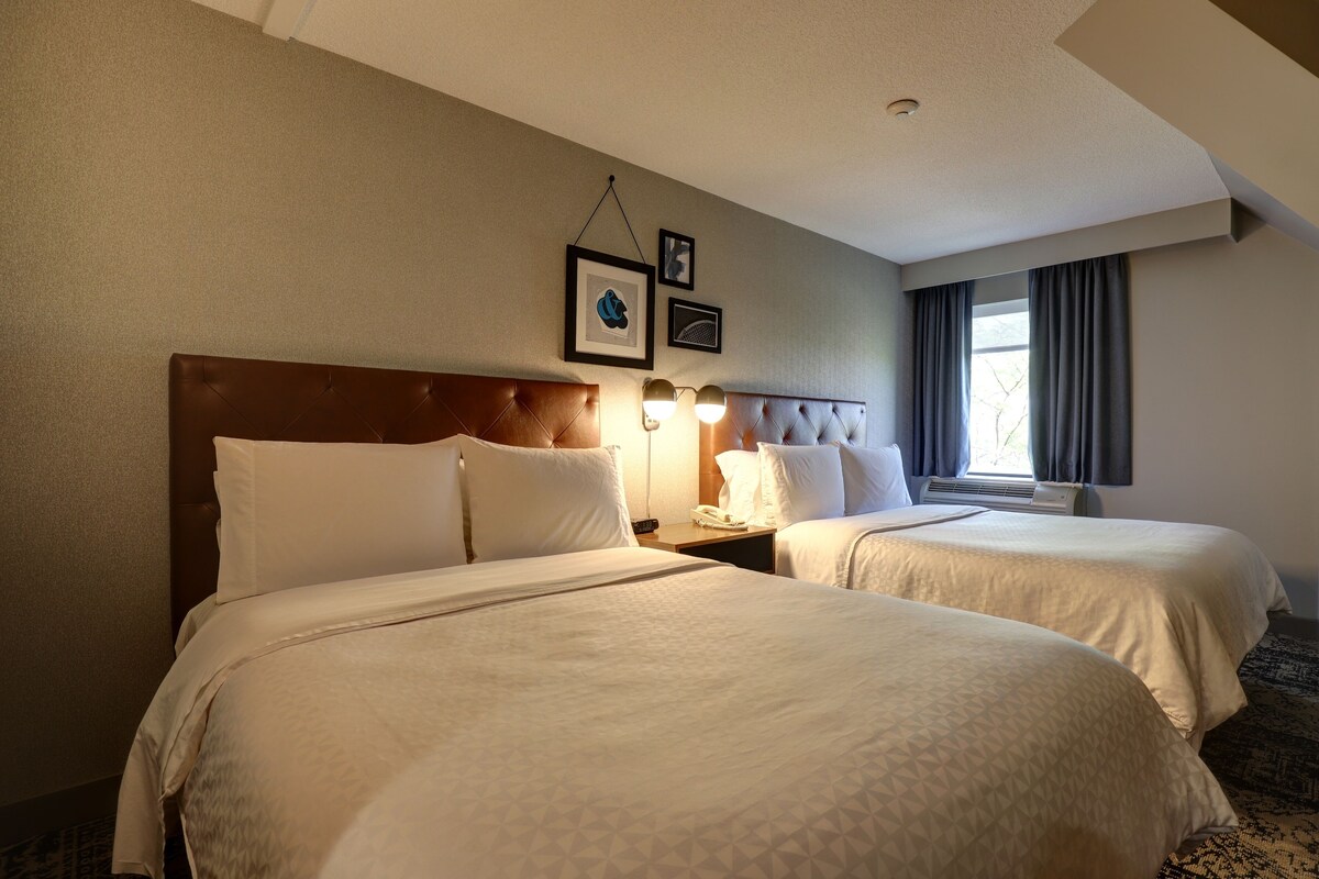 Une chambre d’hôtel avec deux lits, une tête de lit, des tables de chevet, une lampe et une fenêtre avec des rideaux.