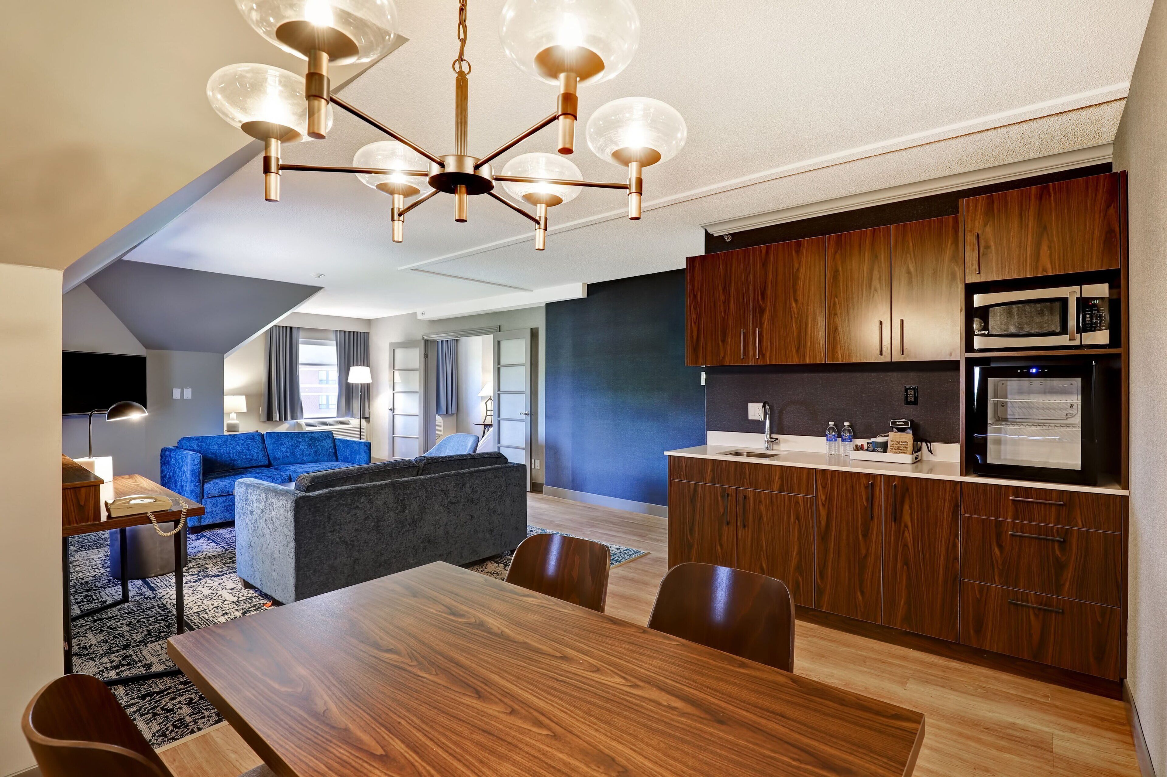 A modern kitchen with wooden cabinets and built-in appliances, a dining area with a wooden table and chairs, and a living area with a blue sofa and a gray armchair.