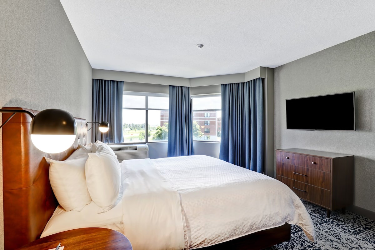 A hotel room with a large bed, a flat-screen TV, a wooden dresser, and a window with blue curtains.