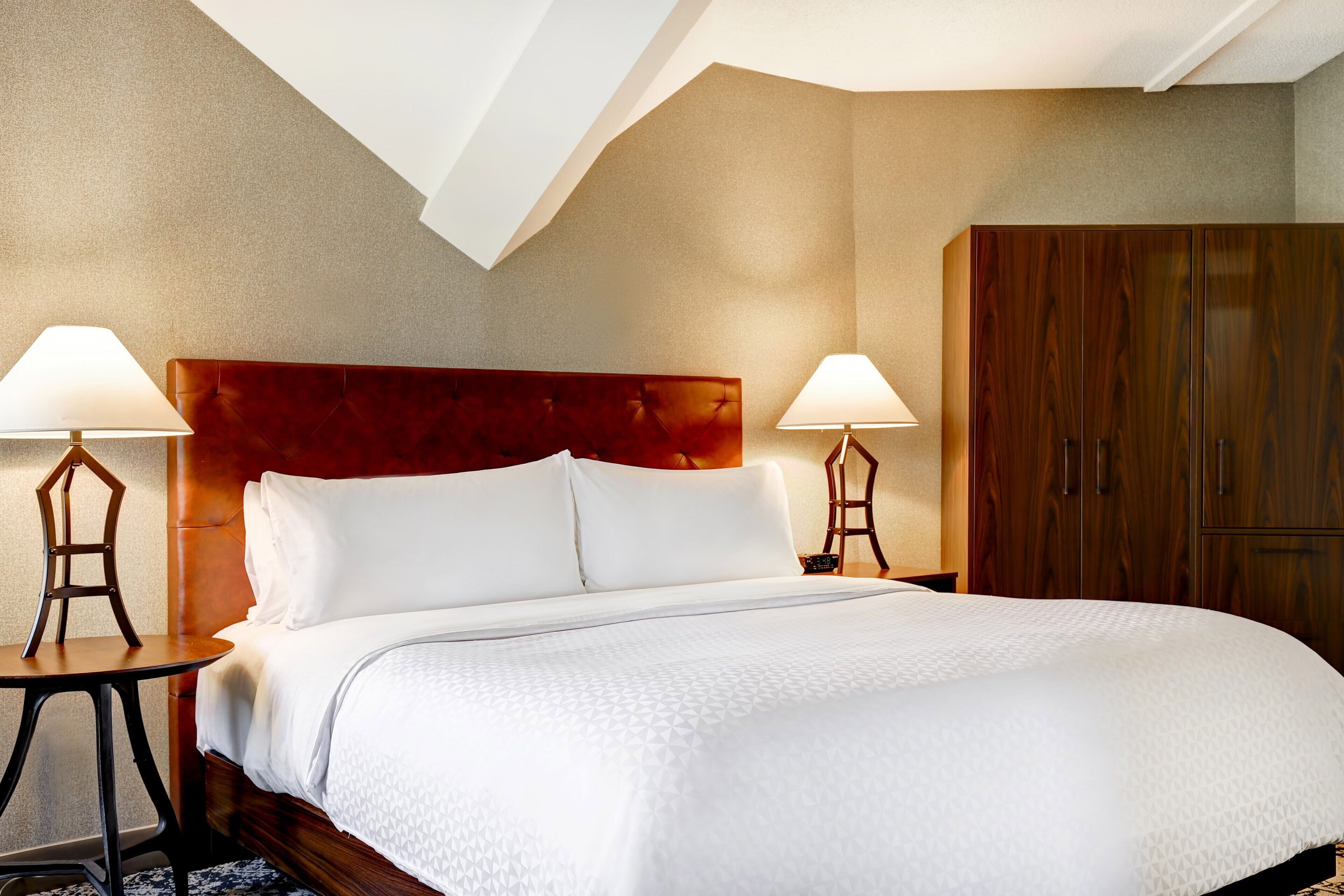 A hotel room with a large bed, two bedside tables with lamps, a wooden wardrobe, and a sloped ceiling.