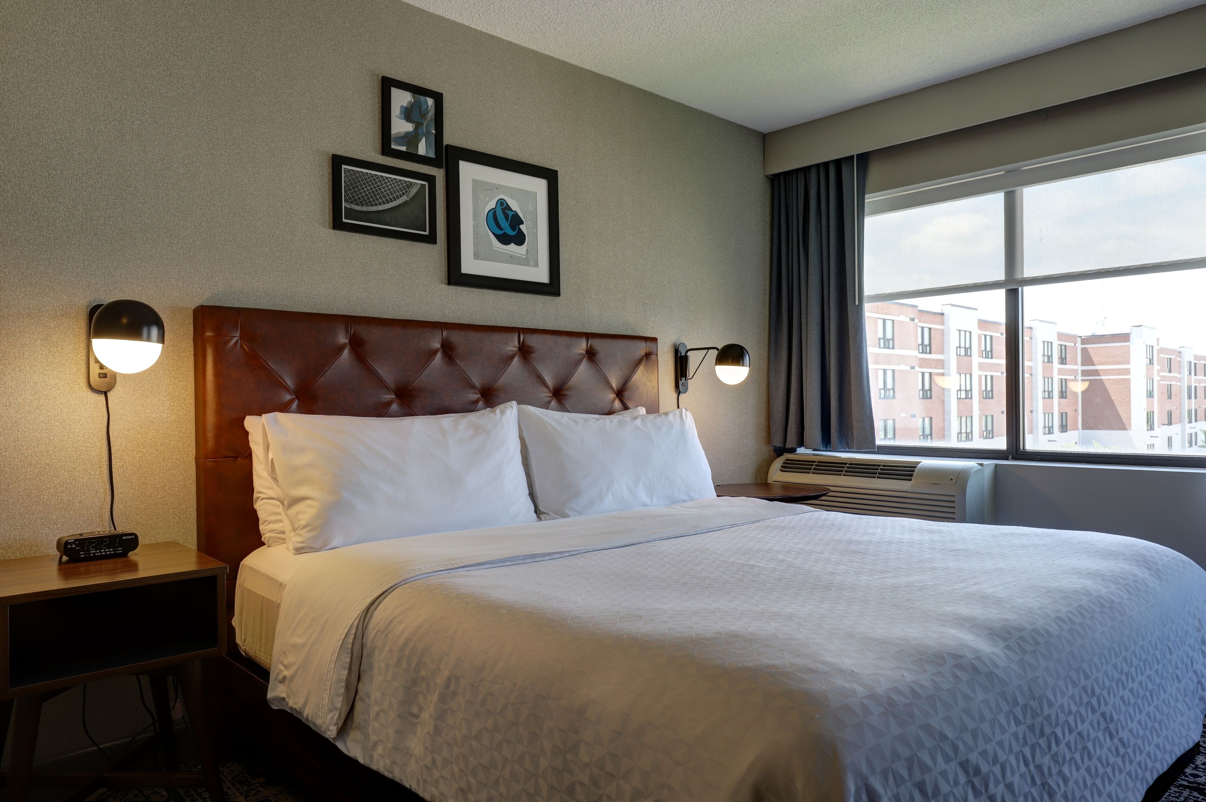 A hotel room with a large bed, two bedside tables, a wall-mounted TV, framed artwork, and a window with a view of buildings.
