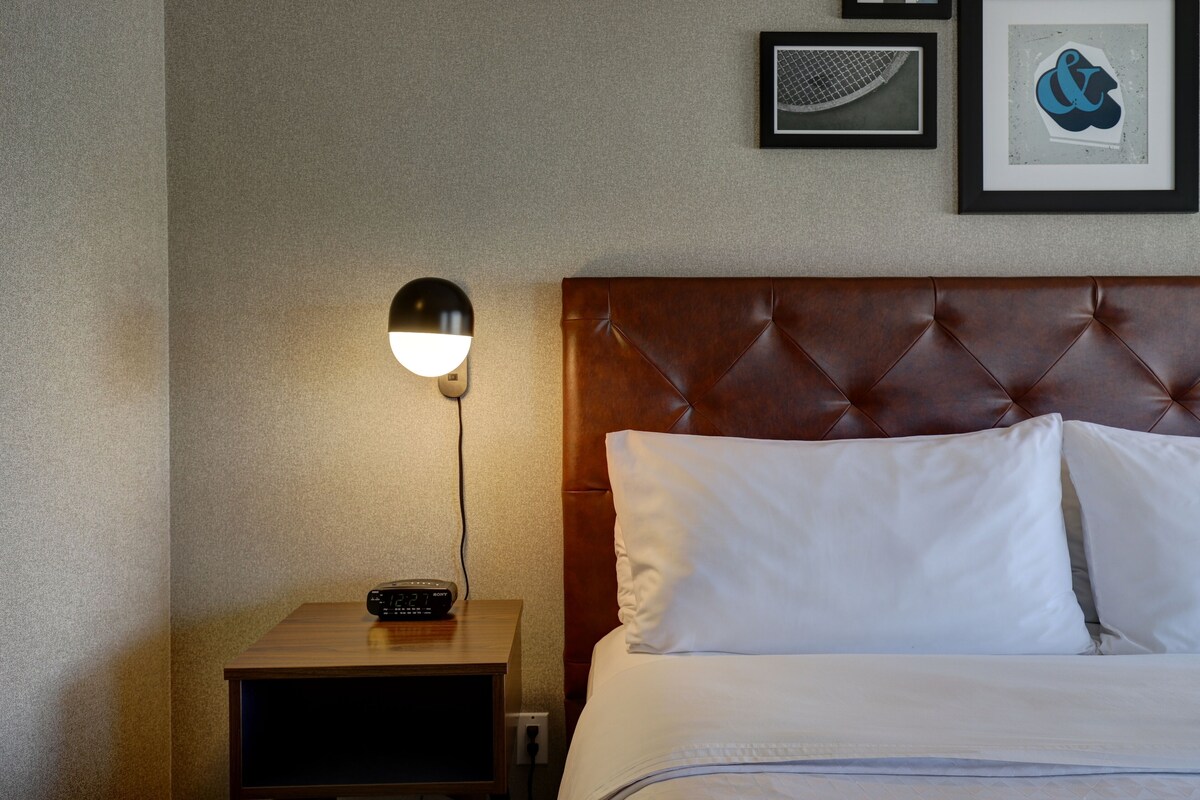 Une chambre d’hôtel avec une tête de lit en bois, une table de chevet avec une lampe et des œuvres d’art encadrées accrochées au mur.