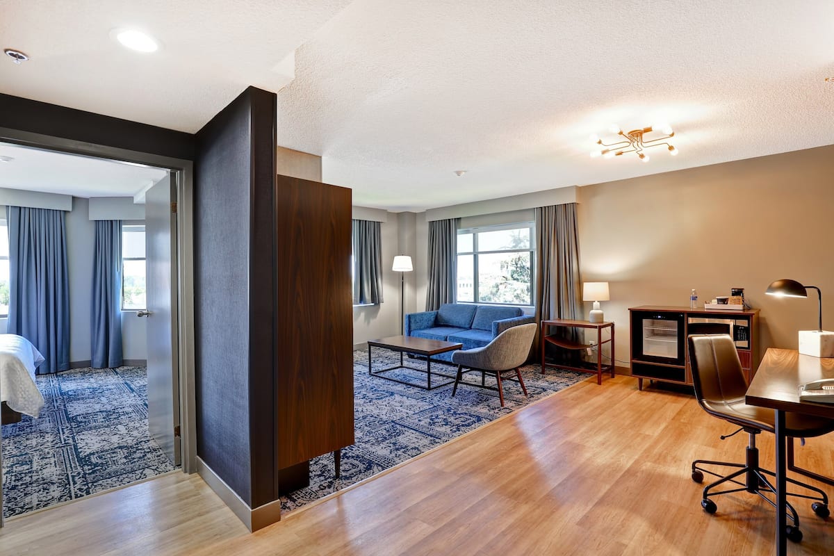 A hotel room with a wooden floor, a blue patterned rug, a sofa, a desk with a chair, and a minibar.