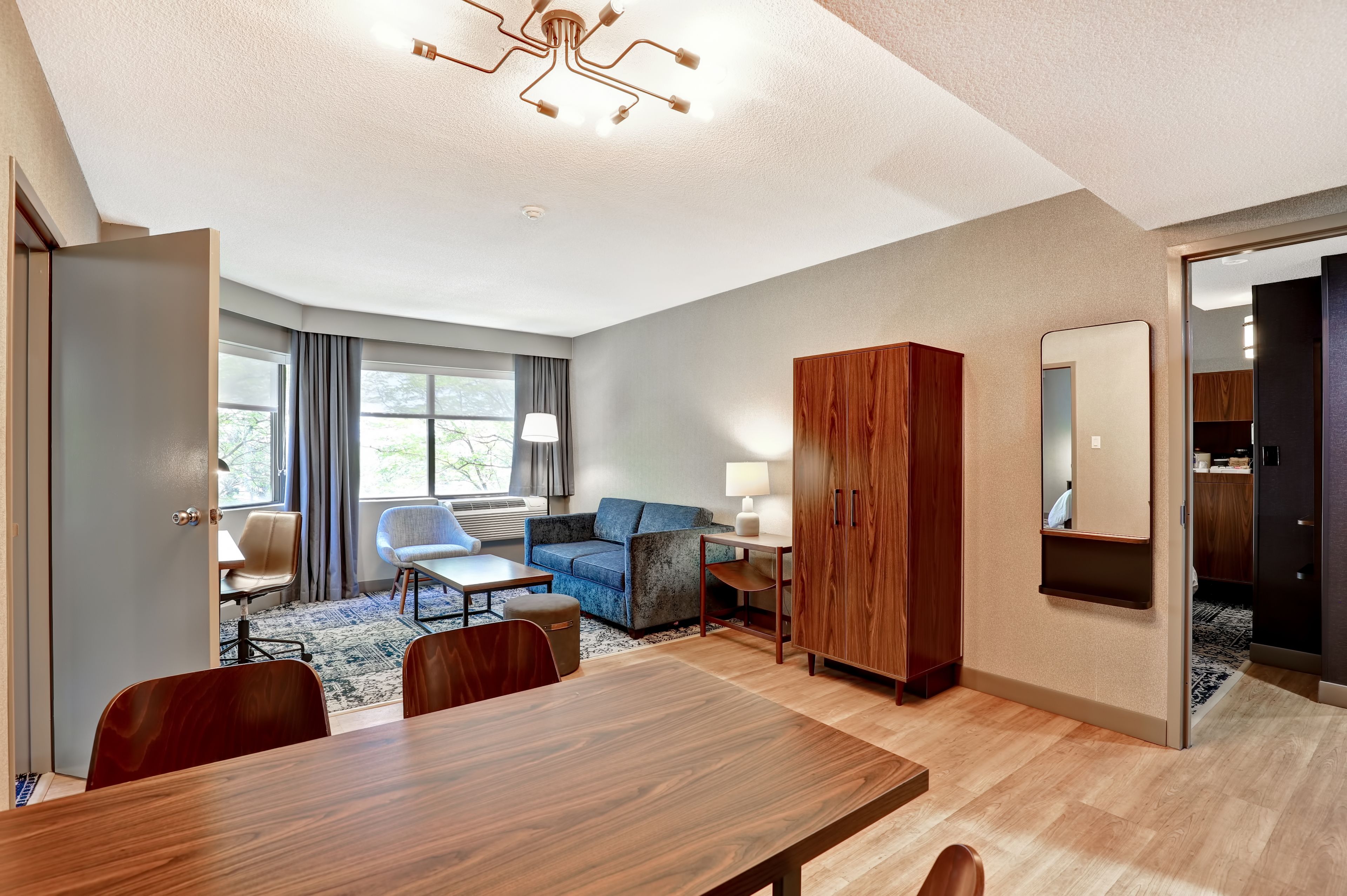 Una habitación de hotel moderna con una mesa de comedor de madera, un sofá azul, un armario de madera y un espejo grande.