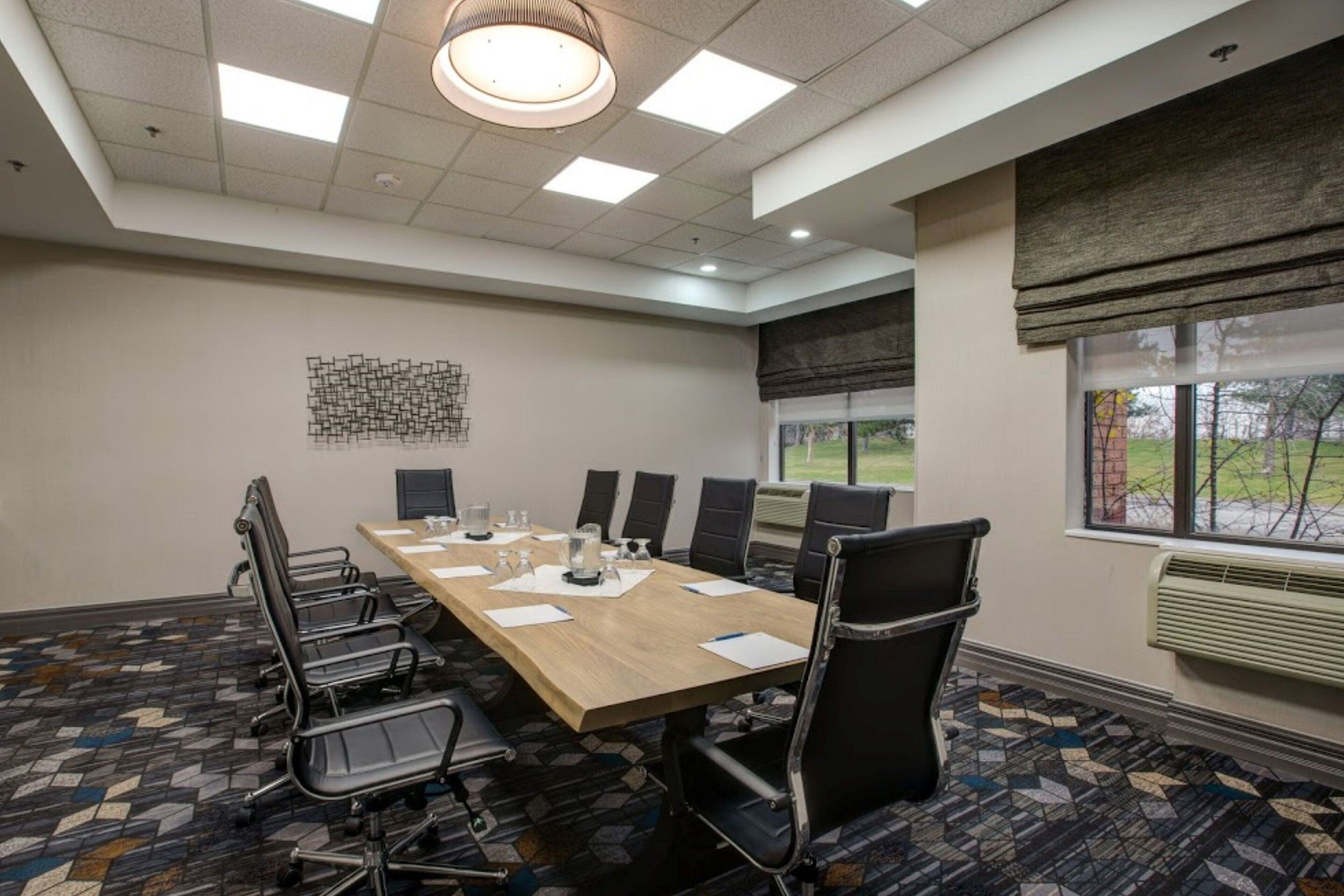 A conference room with a long table, chairs, a patterned carpet, a ceiling light, and a window with blinds.