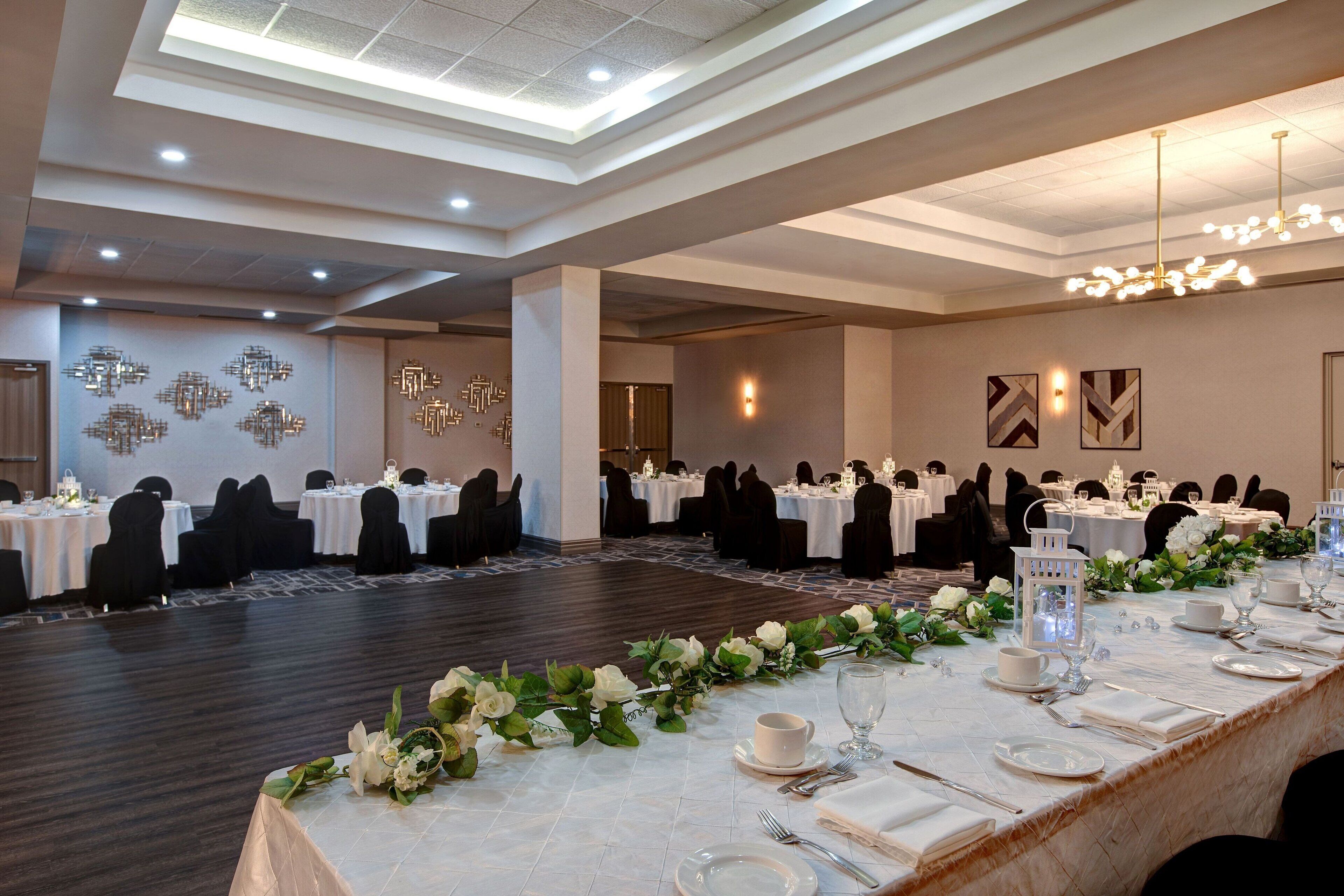 A banquet hall with round tables set for a formal event, featuring a floral centerpiece, white tablecloths, and a chandelier.