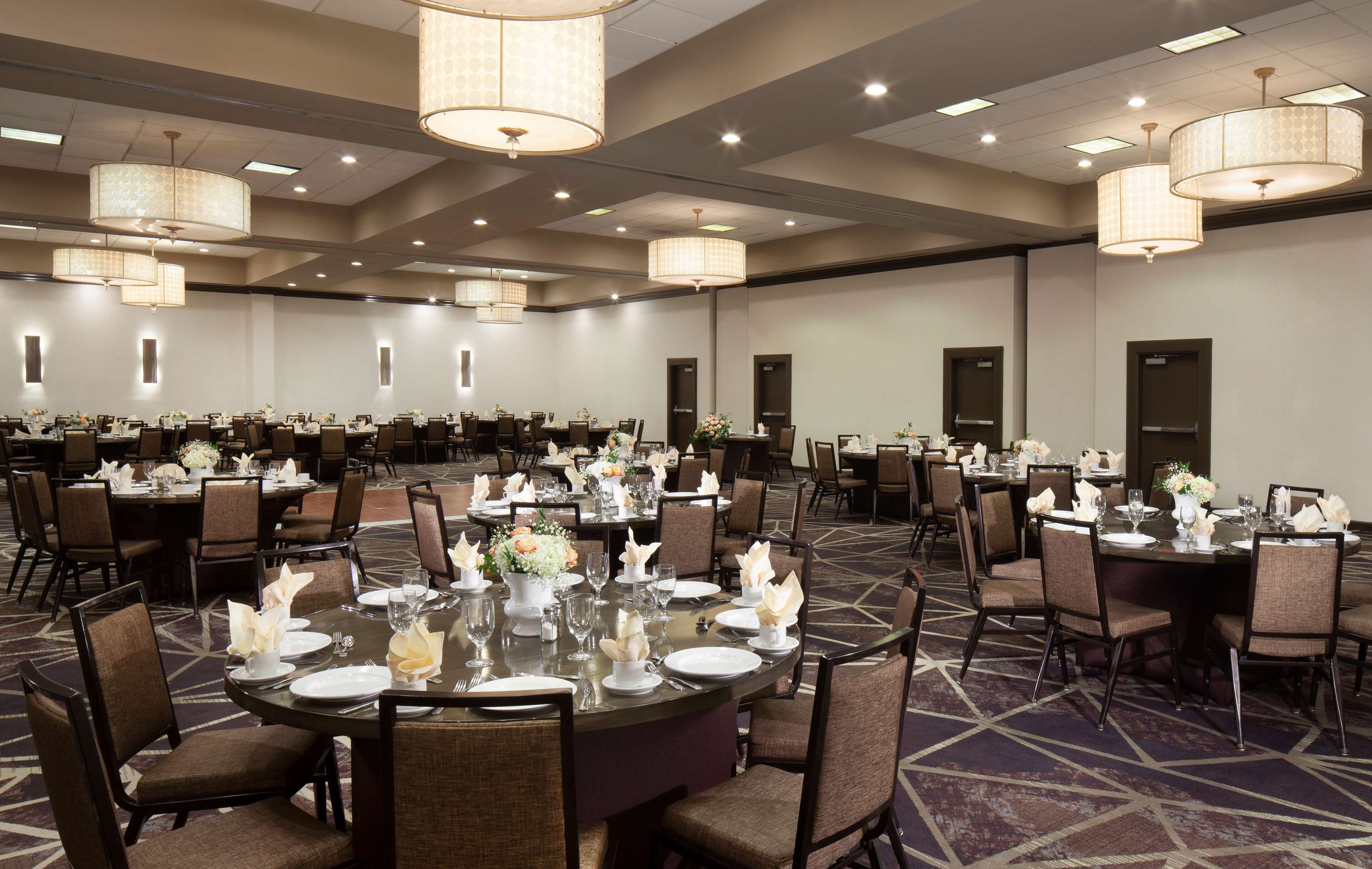 A large conference room with multiple round tables set for dining, equipped with chairs and tables, and decorated with flowers.