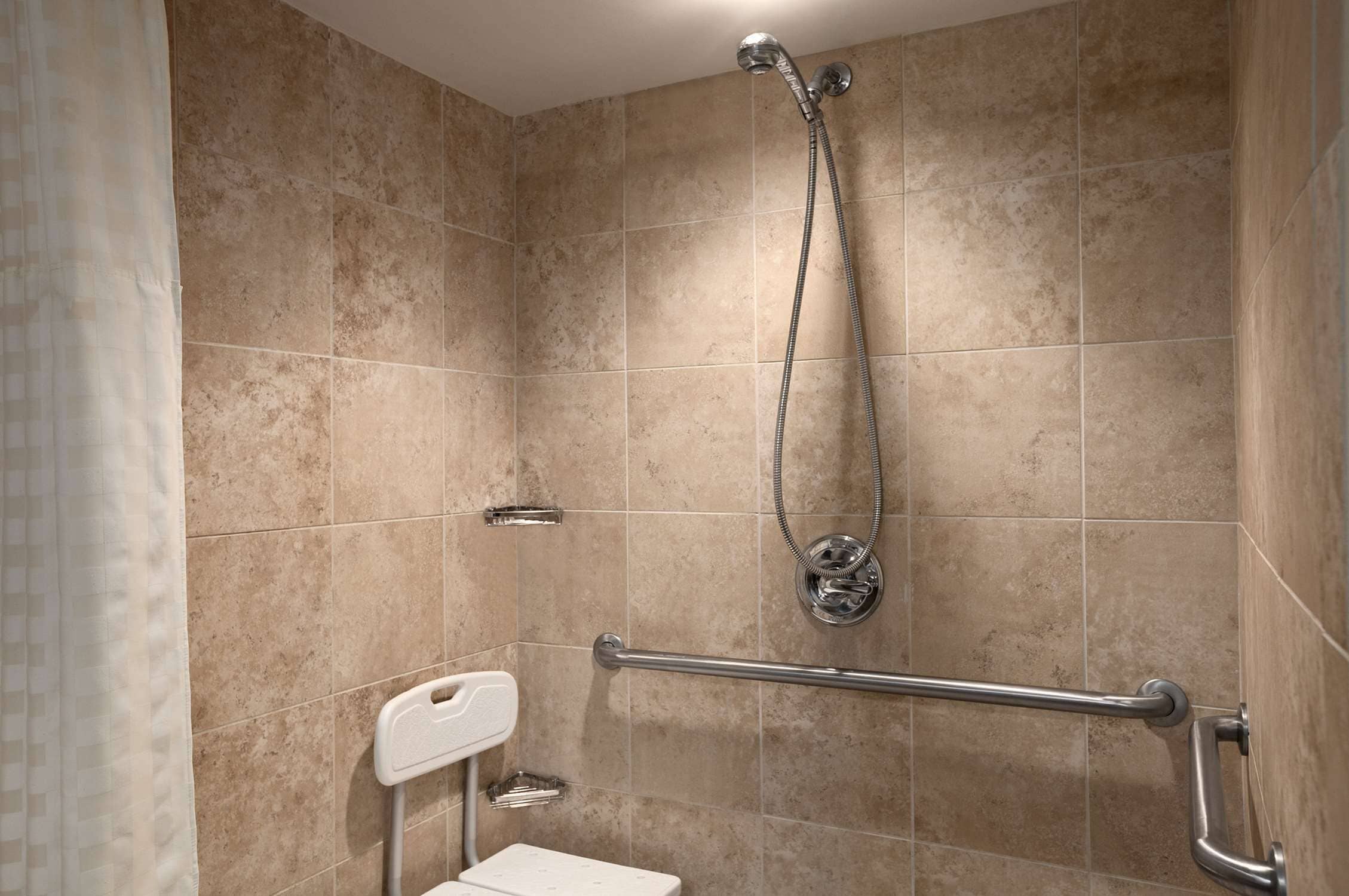 Un baño con azulejos color beige, ducha con cabezal de ducha manual, barra de apoyo y inodoro blanco.