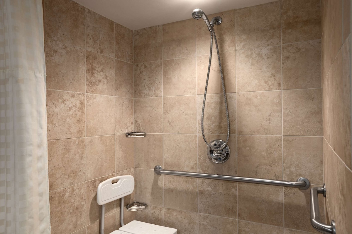 Une salle de bain carrelée de carreaux beiges, une douche avec un pommeau de douche amovible, une barre d’appui et des toilettes blanches.
