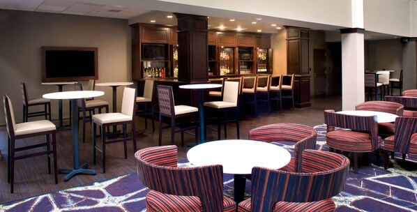 A hotel lobby with multiple seating areas, including round tables and chairs, a bar area with a television, and a carpeted floor.