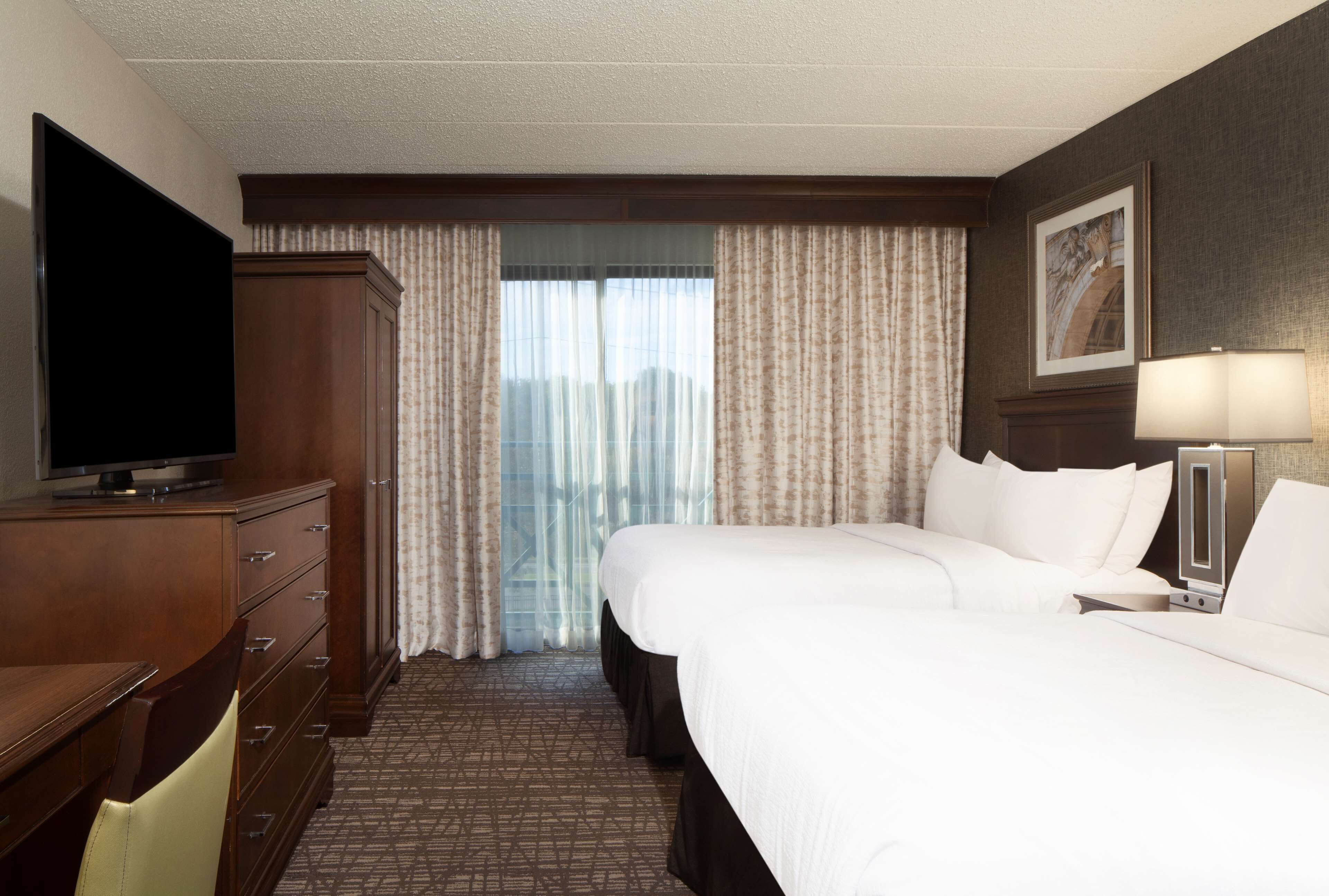 A hotel room with a large bed, a TV on a wooden cabinet, a desk, and a window with curtains.