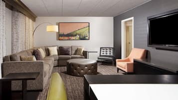 A modern hotel room with a sectional sofa, a round ottoman, a single armchair, a flat-screen TV, and a large painting on the wall.