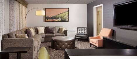A modern hotel room with a sectional sofa, a round ottoman, a single armchair, a flat-screen TV, and a large painting on the wall.