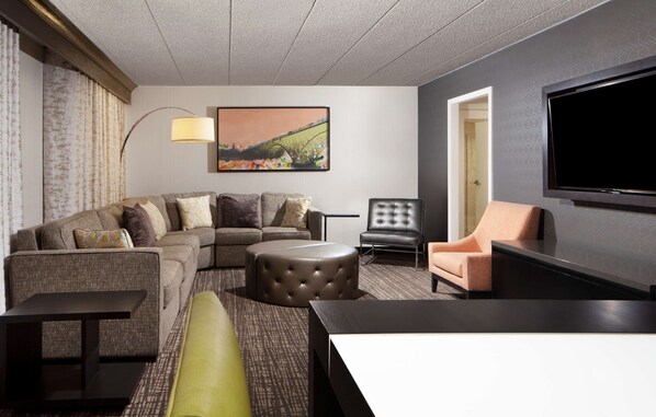 A modern hotel room with a sectional sofa, a round ottoman, a single armchair, a flat-screen TV, and a large painting on the wall.