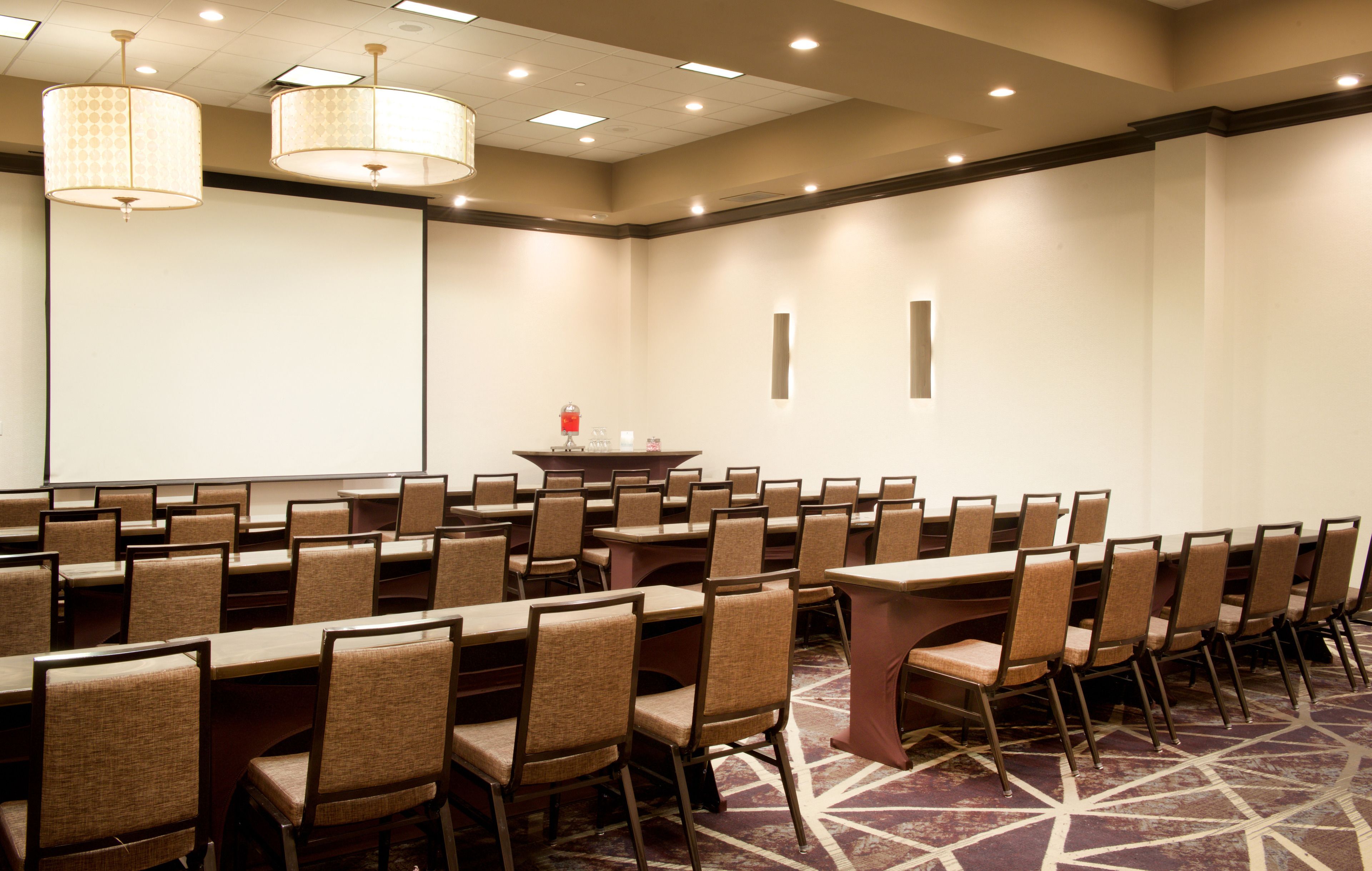 A conference room with rows of tables and chairs, a projector screen, and a podium with a red object on it.