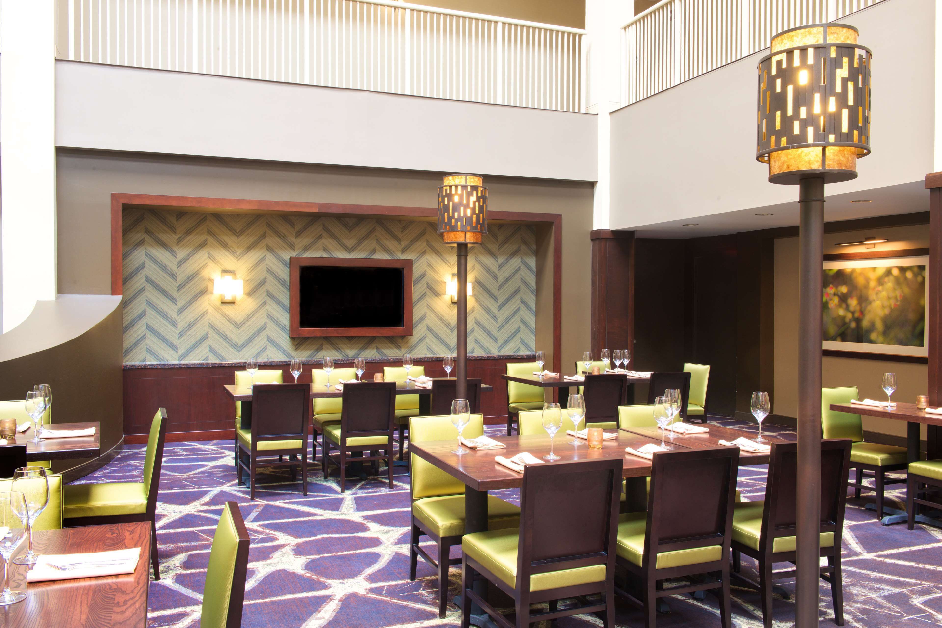 A hotel dining area with multiple tables and chairs, a television mounted on the wall, and a patterned carpet.