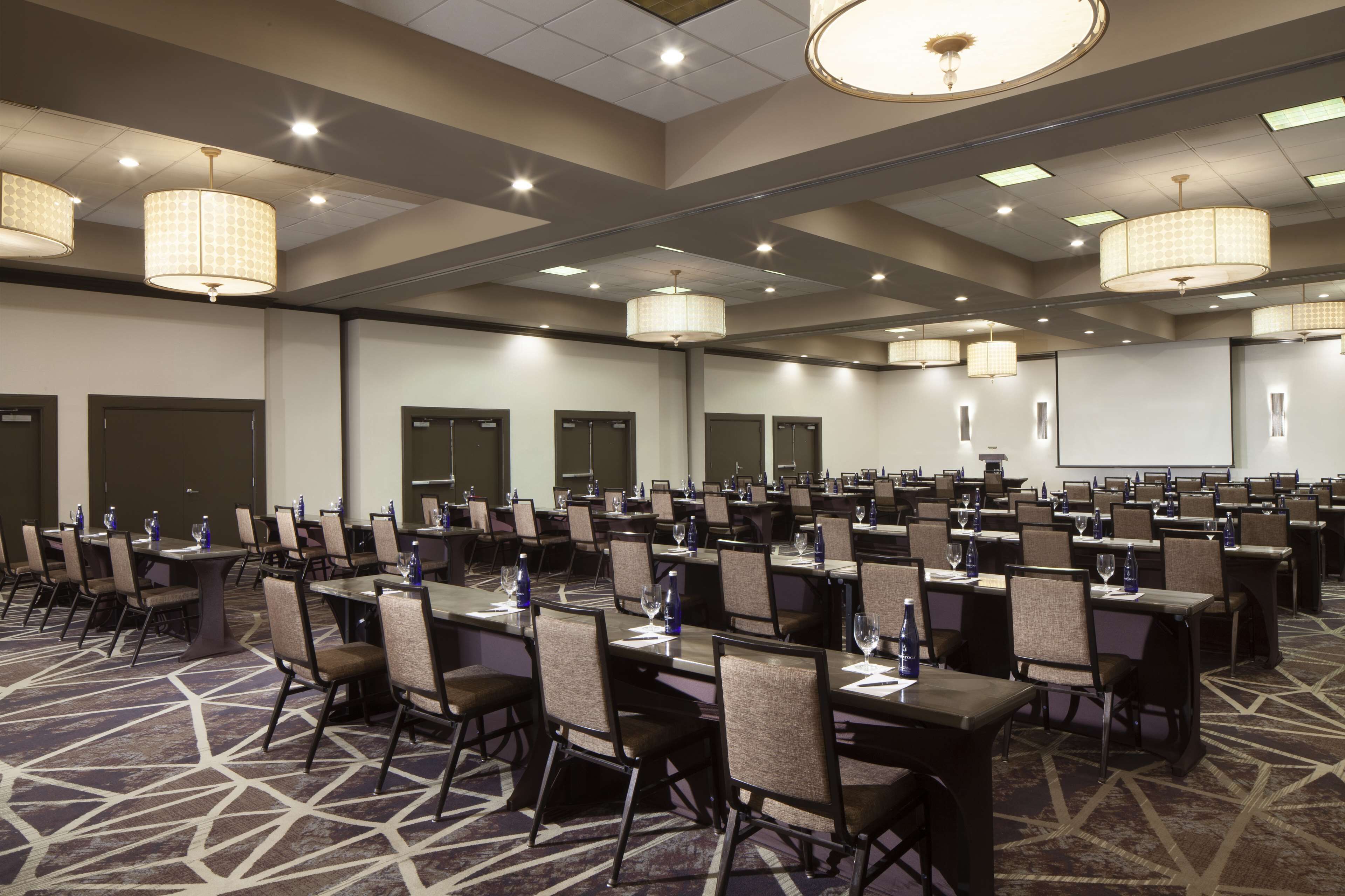 A conference room with multiple tables and chairs, equipped with water bottles and papers, and a projector screen on the wall.