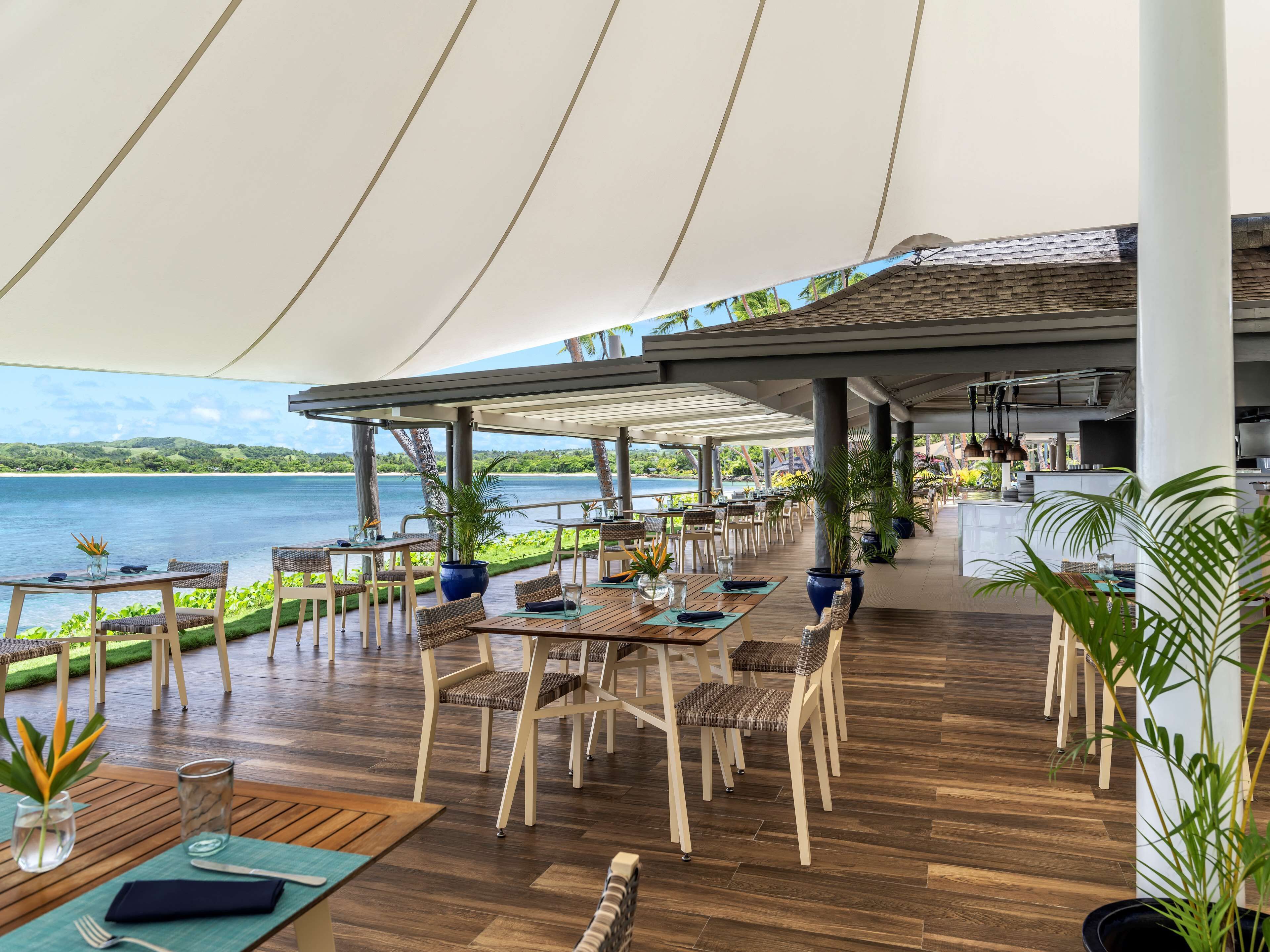 Un área de comedor al aire libre con mesas y sillas, vista al mar y un paseo cubierto.