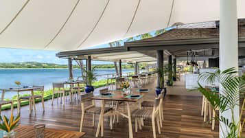 Un área de comedor al aire libre con mesas y sillas, vista al mar y un paseo cubierto.