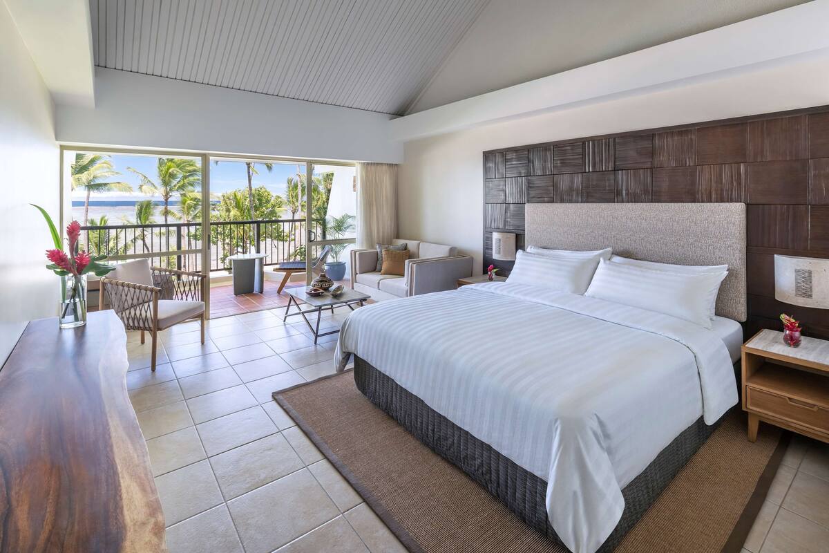A modern hotel room with a large bed, a balcony with a view of the beach, a dining area, and a wooden desk.