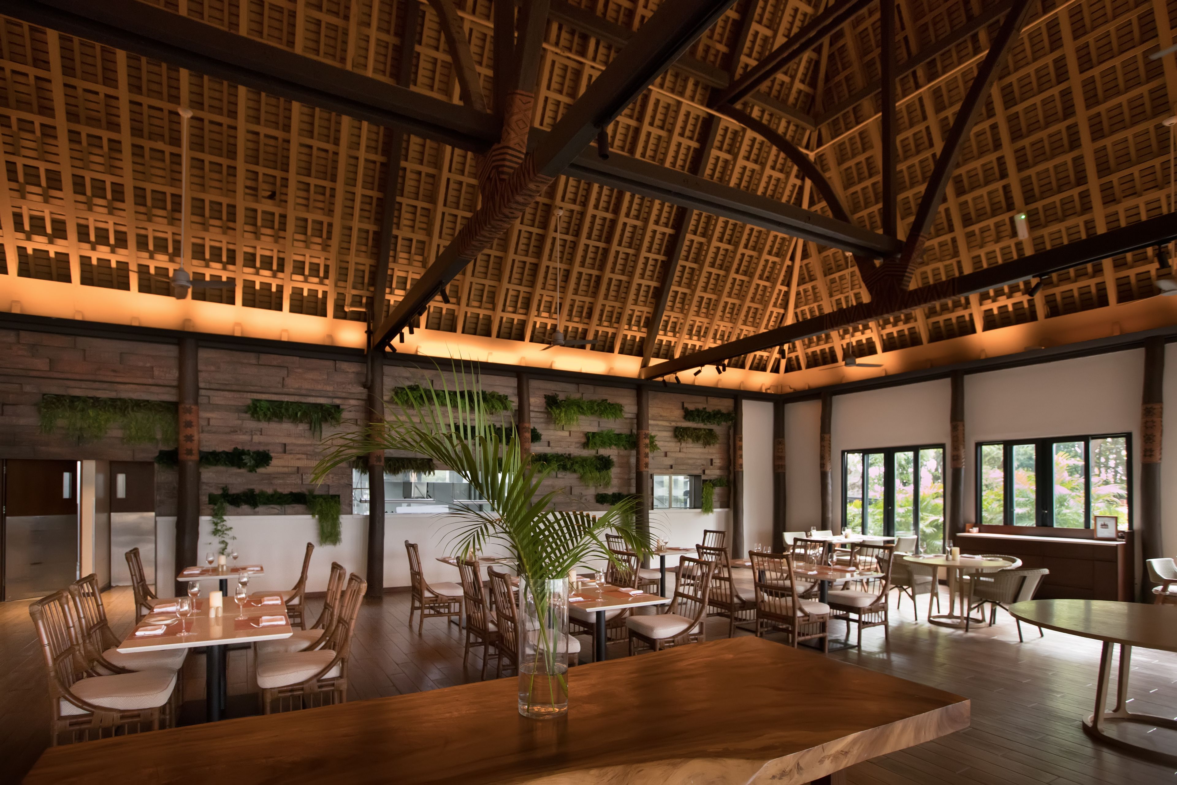 Un restaurante espacioso con techos altos, vigas de madera y una decoración interior moderna. Las mesas están dispuestas con manteles blancos y velas.