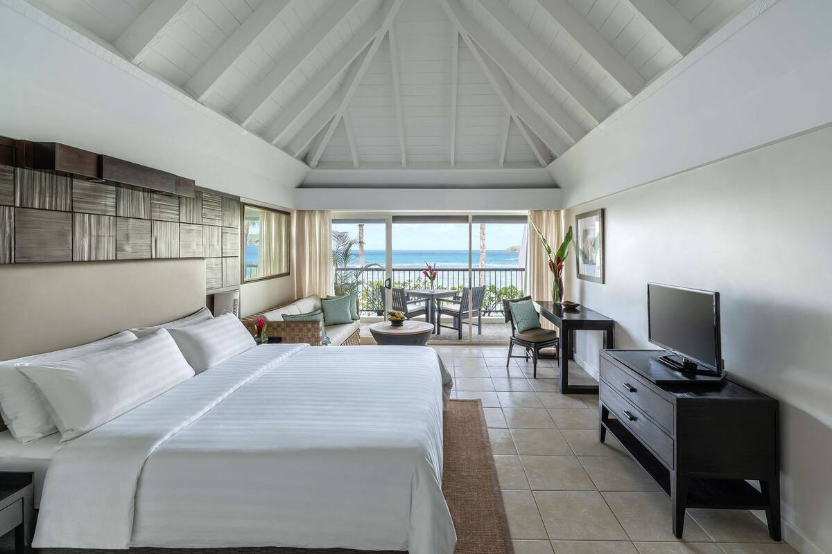 A hotel room with a large bed, a desk with a TV, a balcony with outdoor seating, and a view of the ocean.