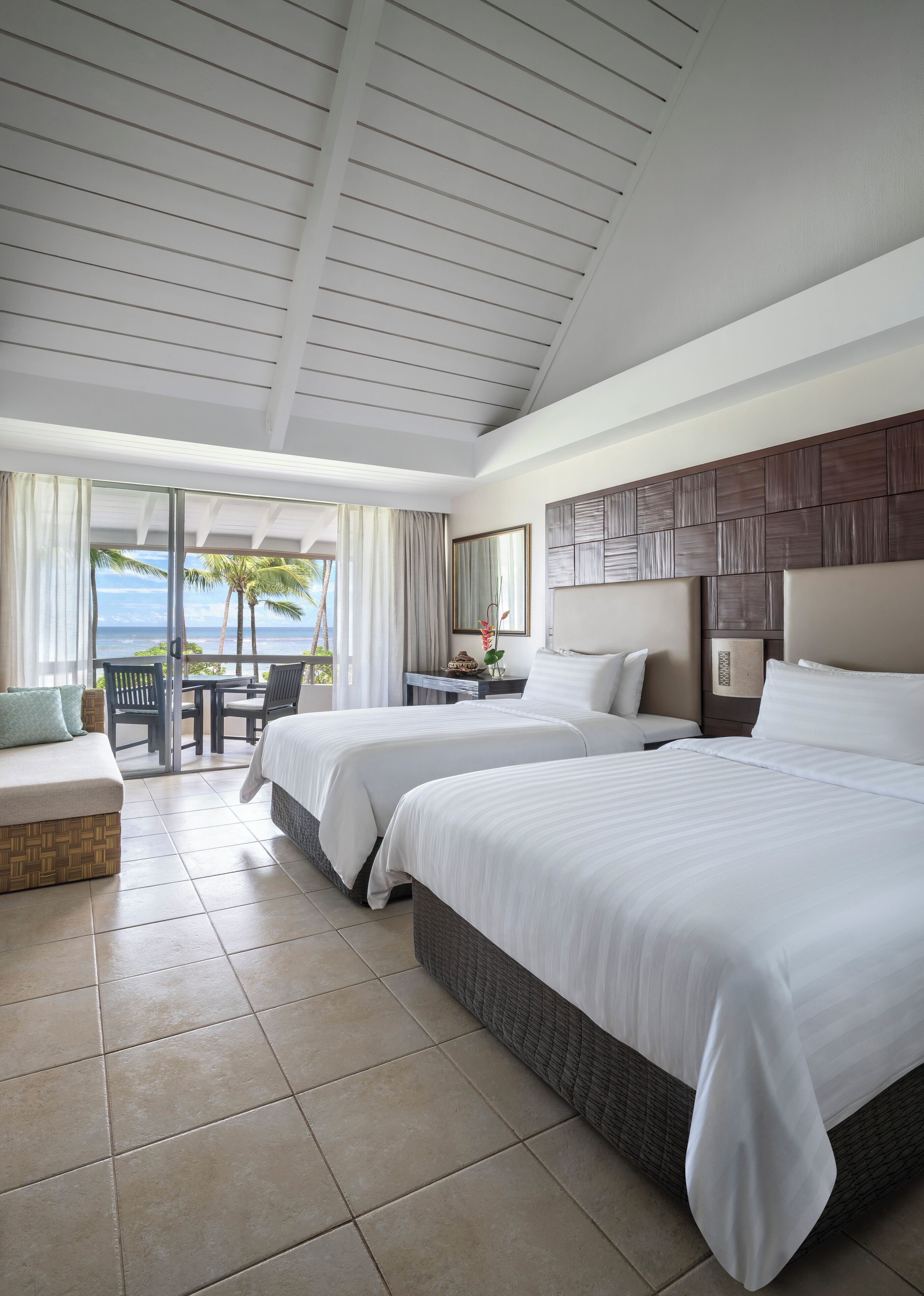 A hotel room with two beds, a balcony with a view of the ocean, and a dining area with a table and chairs.