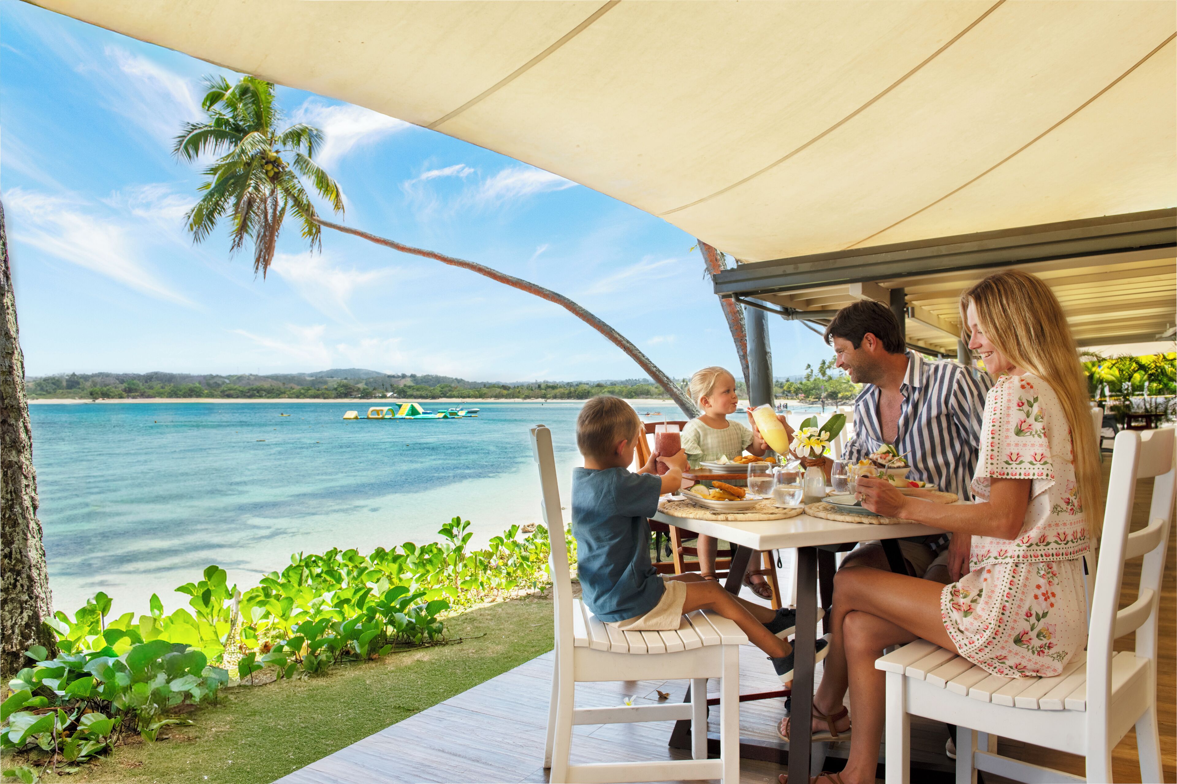 A family dining at an outdoor table with a view of the ocean and palm trees.