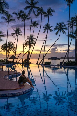 Zona junto a la piscina con palmeras, sillas de descanso y vista al atardecer sobre el agua.