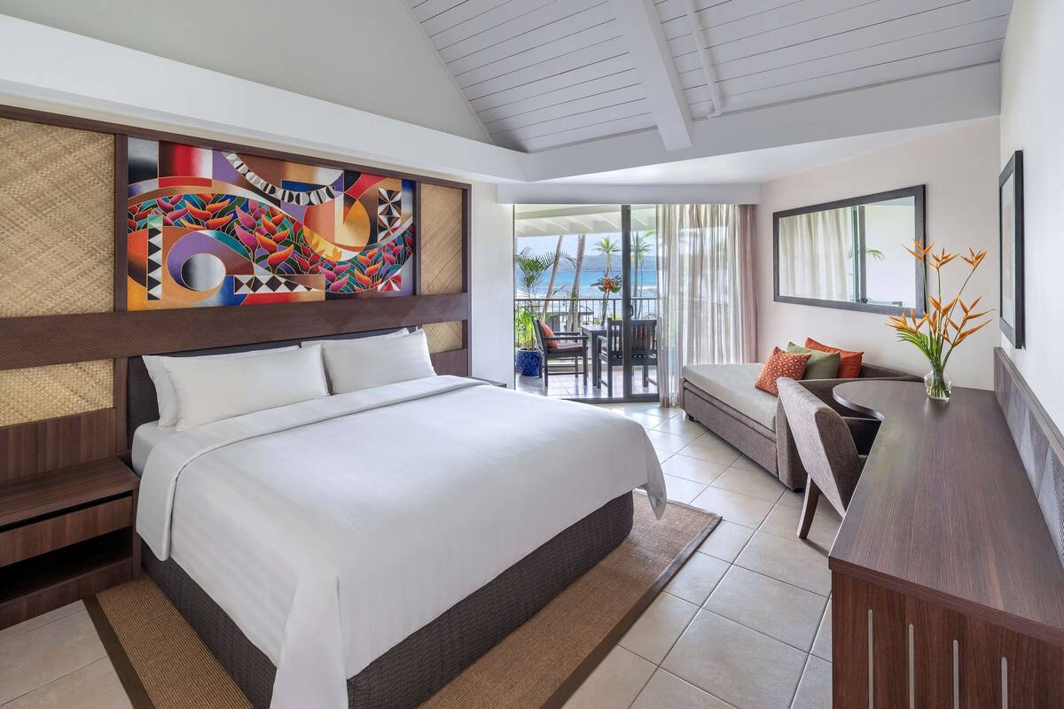 A hotel room with a large bed, a desk, a balcony with a view of the ocean, and a colorful abstract painting on the wall.