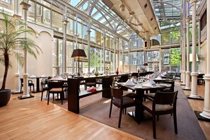 A spacious conservatory with high ceilings and large windows, featuring multiple dining tables set for guests. - Hilton London Euston (London)