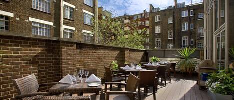An outdoor patio with wicker furniture and tables set up for dining, surrounded by brick buildings.