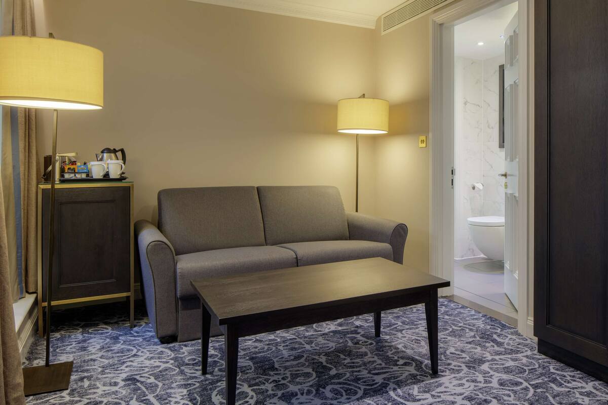 Et hotelværelse med sofa, et lille bord, en lampe, et skab og et badeværelse, der kan ses igennem en åben dør.