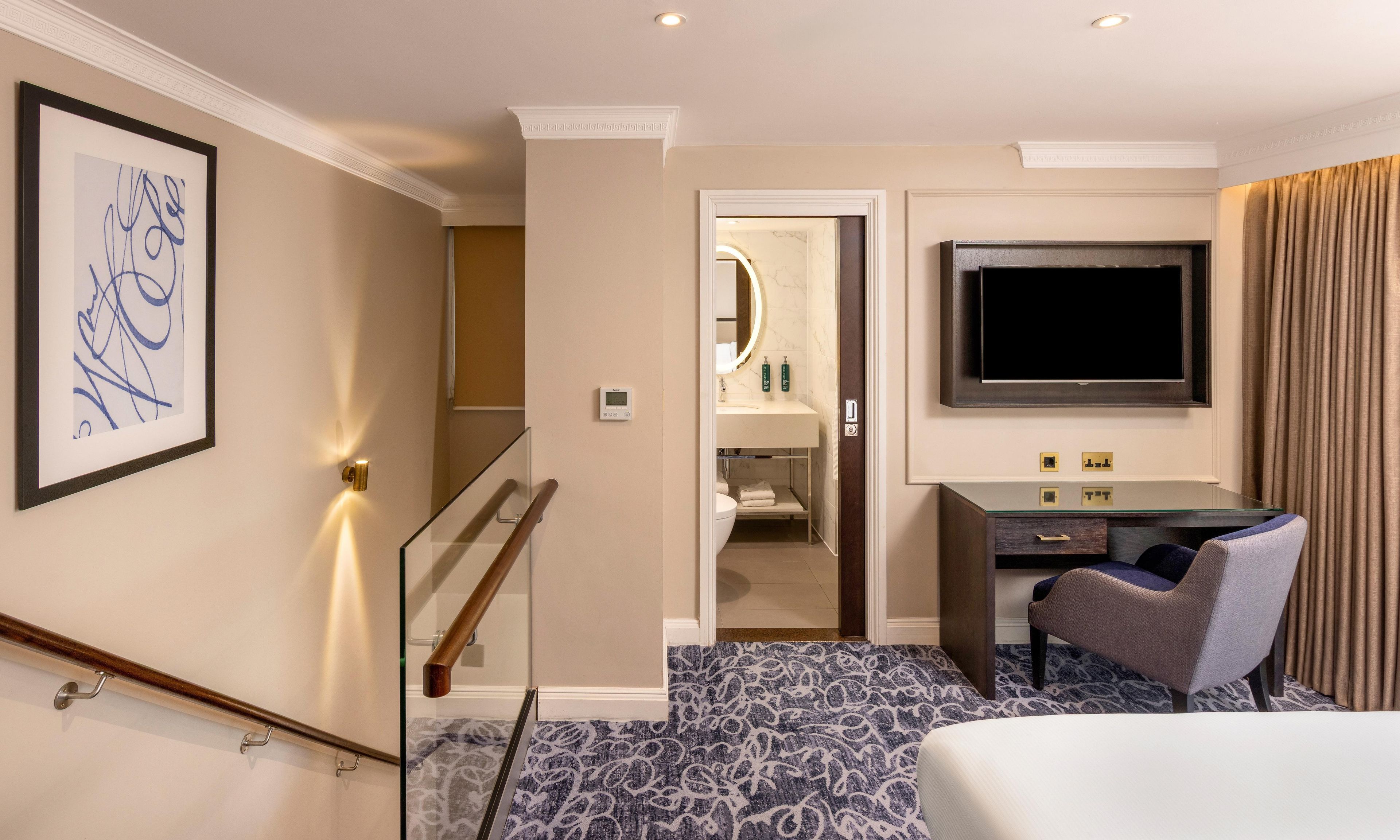 A hotel room with a bed, a desk with a chair, a television, and a bathroom visible through an open door.