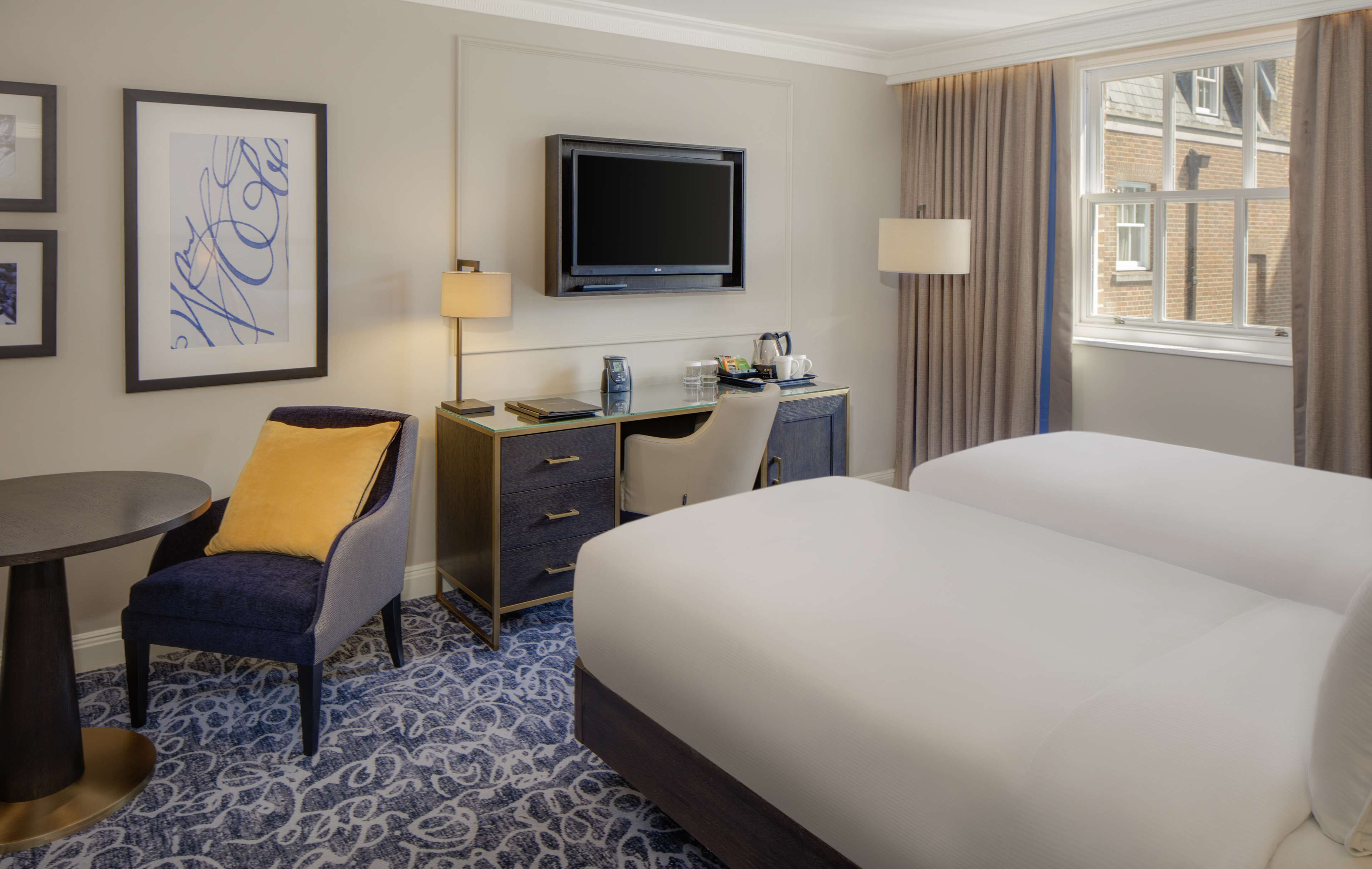 A hotel room with a bed, desk, chair, TV, and artwork on the wall.