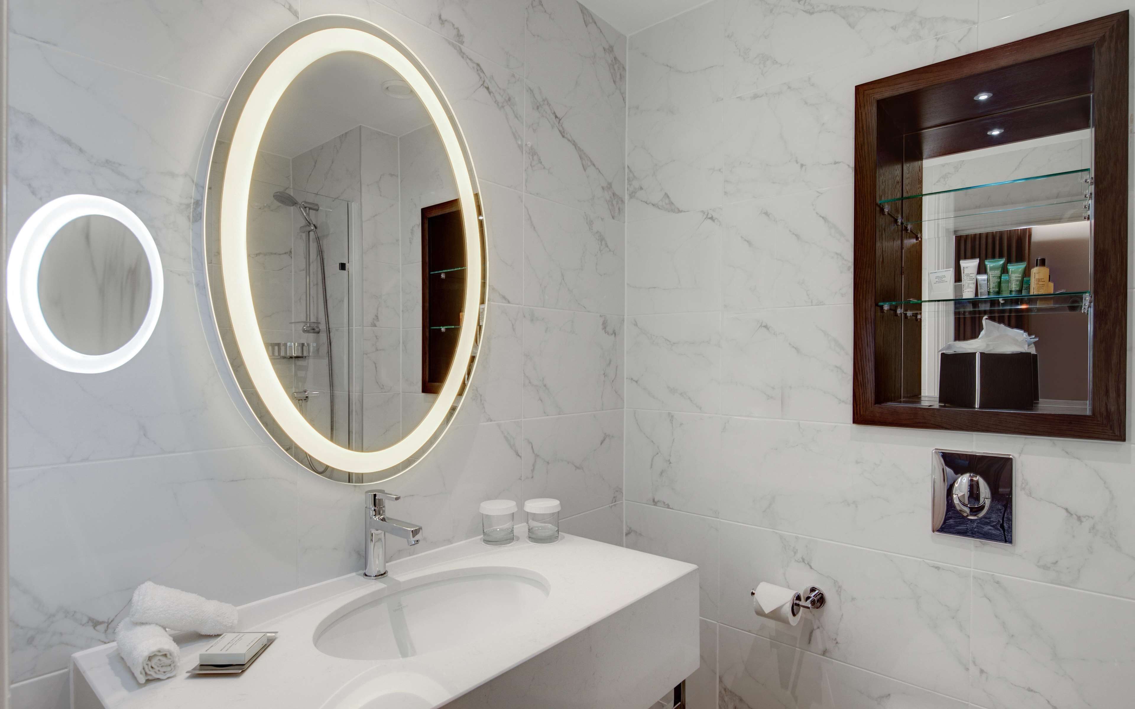 A modern bathroom with a large oval mirror, a white sink, and a glass shower enclosure.