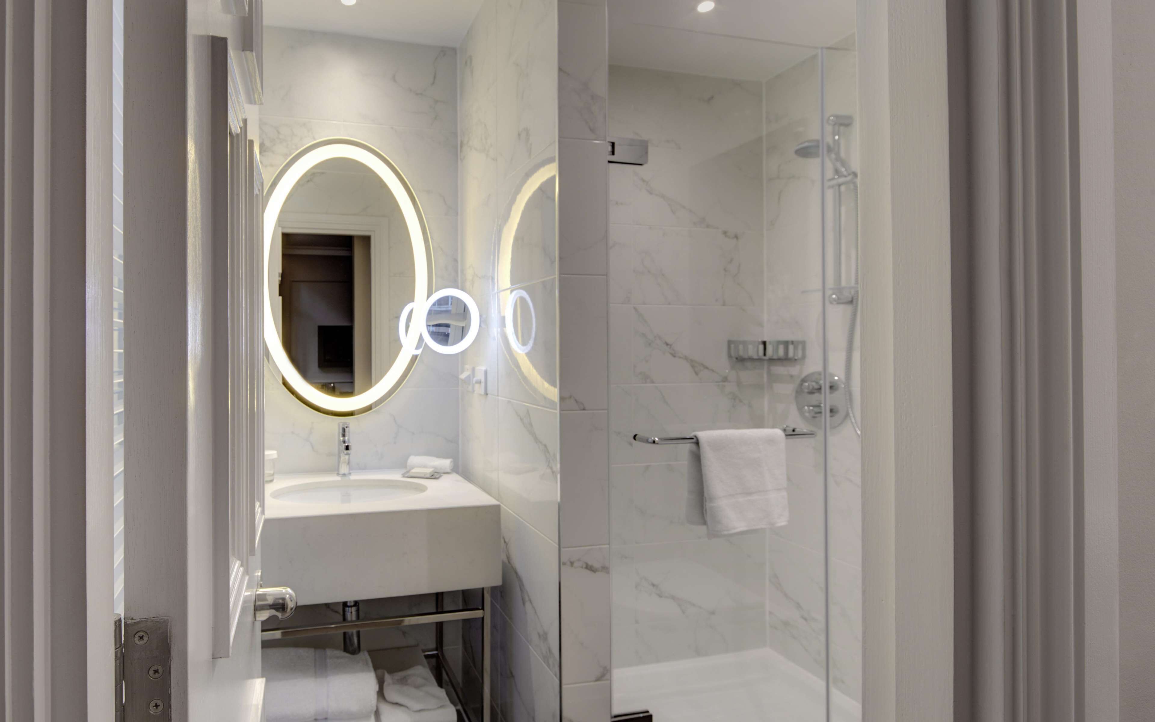 Un baño moderno con lavamanos de mármol, ducha con cerramiento de vidrio y espejo de tocador con iluminación circular.